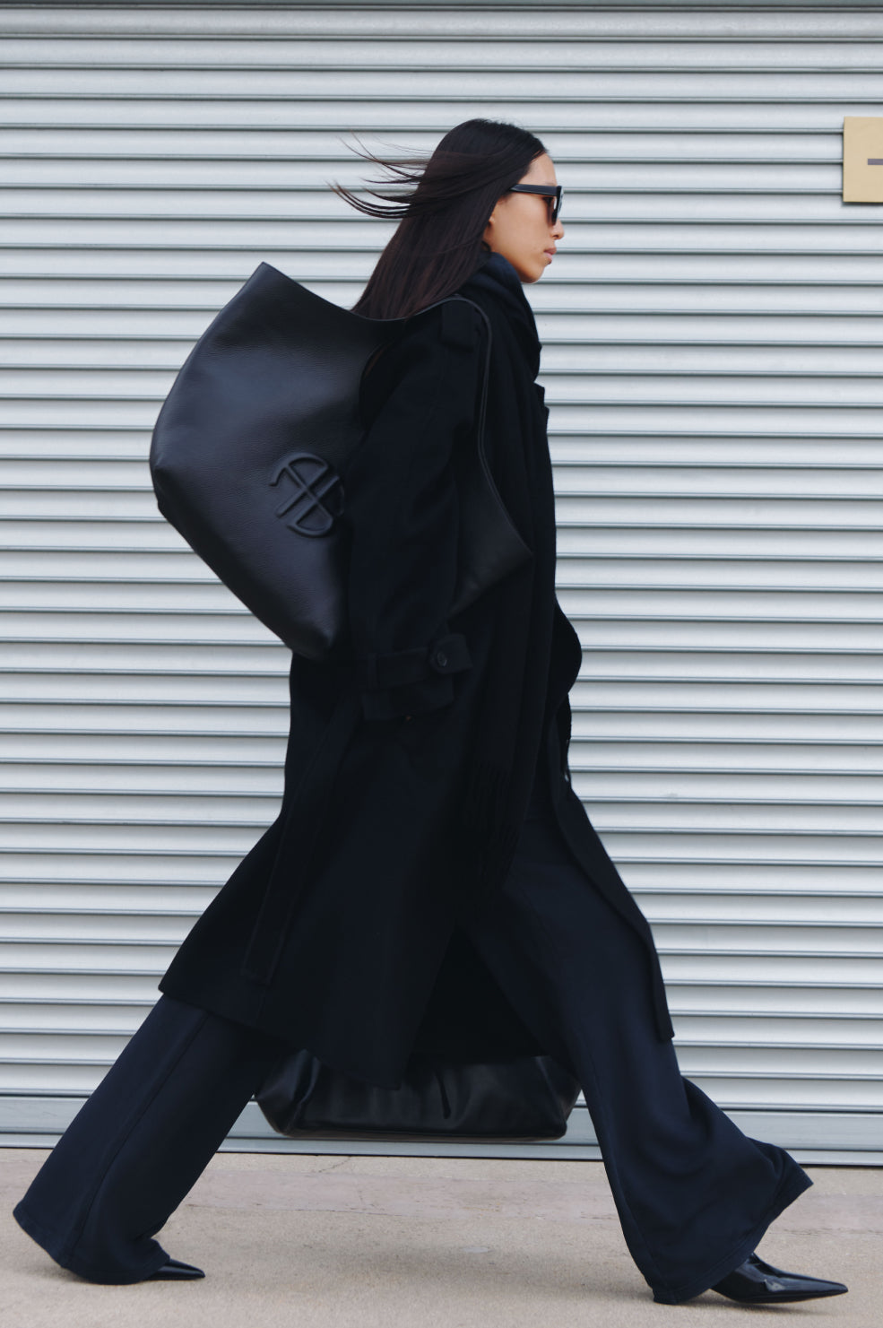 Person wearing a long black coat and carrying a matching black bag against a metallic shutter background