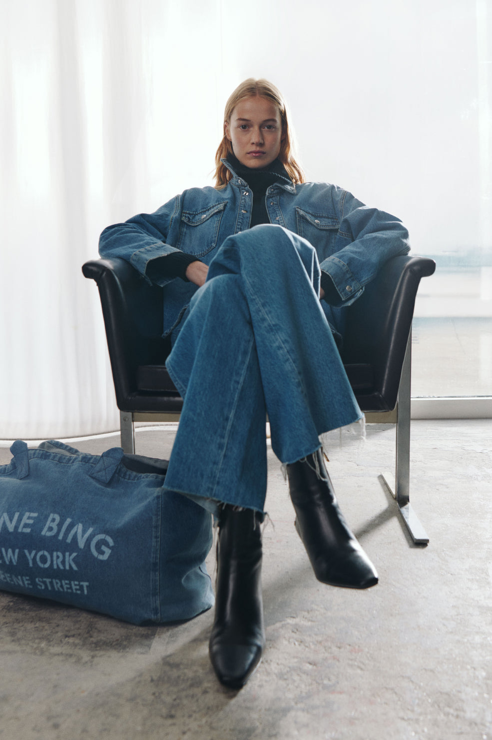 Person wearing a denim outfit sitting in a chair with a blue bag beside them.